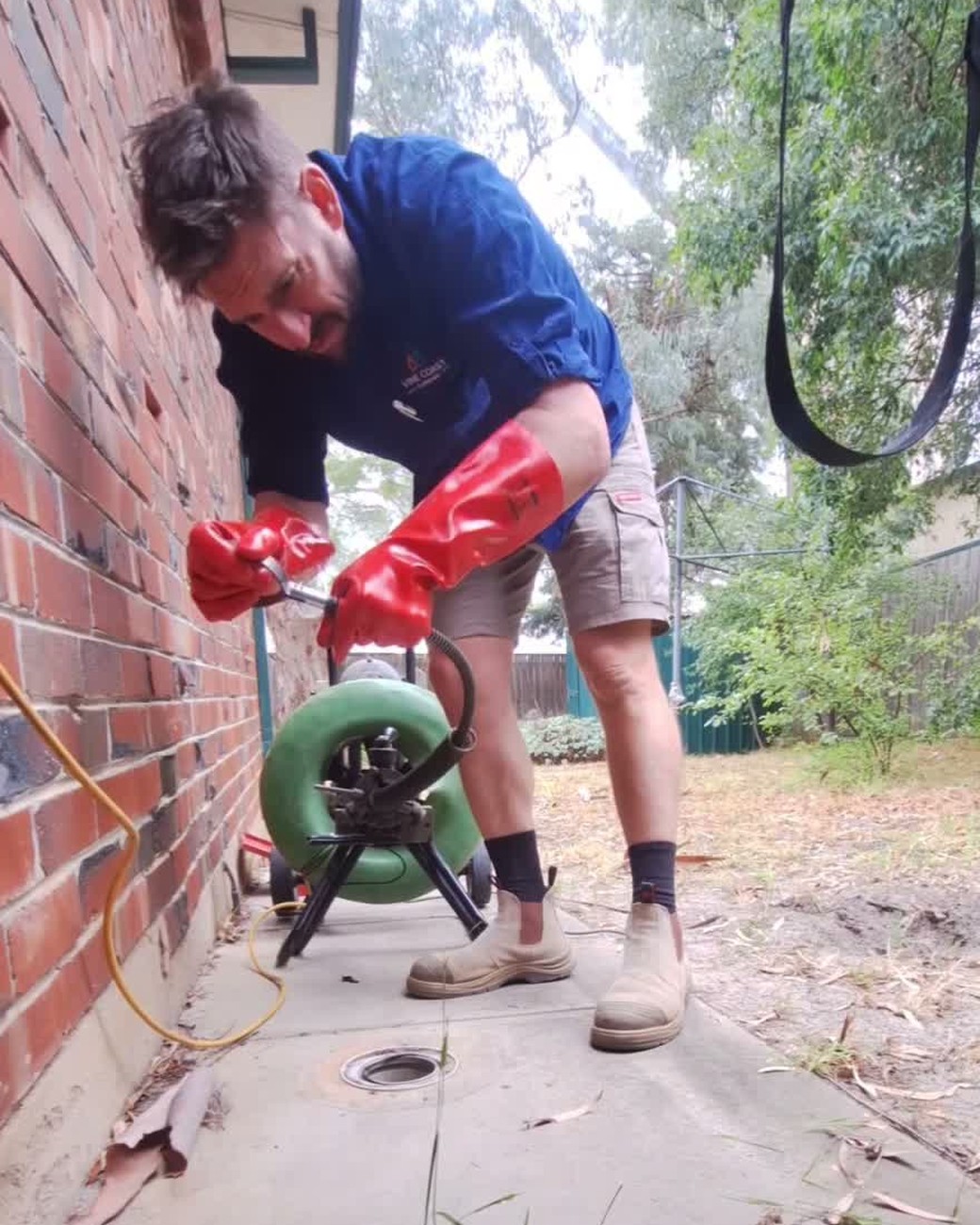 Arthur Lynas in a blue Vine Coast Plumbing shirt and red gloves feeding a drain machine beside a brick house